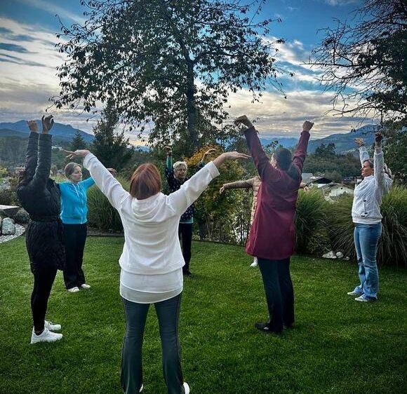 Eine Gruppe von Frauen praktiziert Yoga auf einem Rasen mit Bäumen und Bergen im Hintergrund.