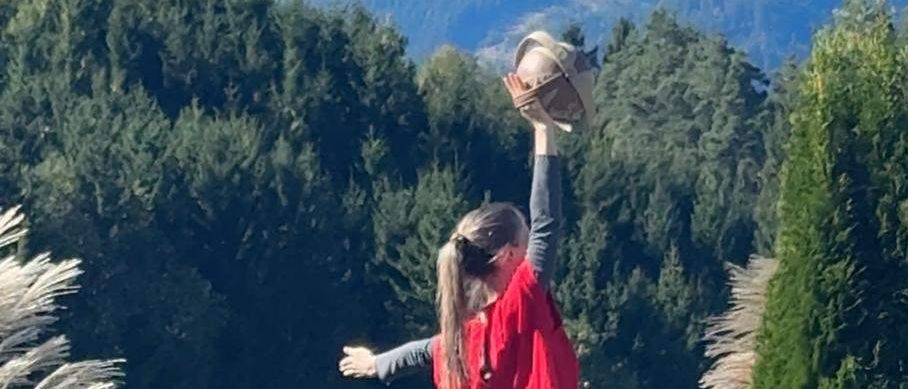 Eine Frau in einem roten Shirt und grauen Hosen steht auf einem abschüssigen Bereich in einem Garten und streckt ihren Arm nach oben. Hinter ihr befindet sich eine Steinmauer, Pflanzen und eine weiße Statue.