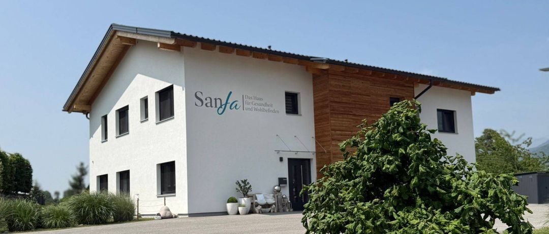 Ein modernes weißes Gebäude mit braunem Dach heißt SanFa. Es gibt mehrere Fenster und eine schwarze Tür. Vor dem Gebäude befindet sich ein Rasenbereich.
