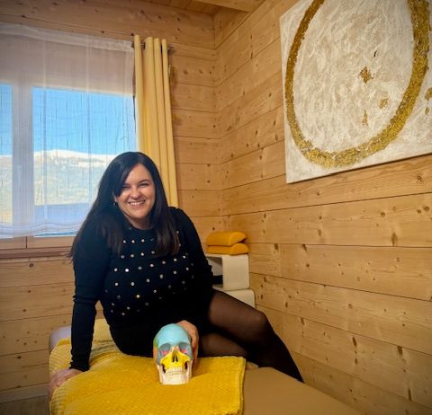 Eine Frau sitzt lächelnd auf einem Bett in einem gemütlichen Holzzimmer, hält einen Schädel und schaut durch ein Fenster. An der Wand hängt ein golden umrandetes Gemälde.