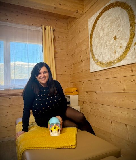 Eine Frau sitzt lächelnd auf einem Bett in einem gemütlichen Holzzimmer, hält einen Schädel und schaut durch ein Fenster. An der Wand hängt ein golden umrandetes Gemälde.