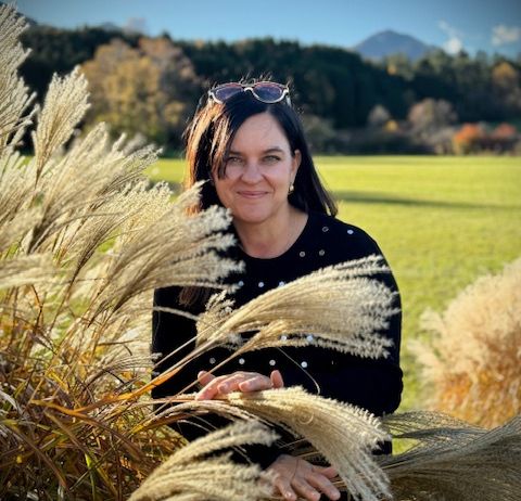 Eine Frau steht in einem Feld aus hohen, goldenen Gräsern, trägt ein schwarzes Shirt und Sonnenbrille auf dem Kopf, mit einem Berg im Hintergrund.