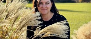 Eine Frau steht in einem Feld aus hohen, goldenen Gräsern, trägt ein schwarzes Shirt und Sonnenbrille auf dem Kopf, mit einem Berg im Hintergrund.