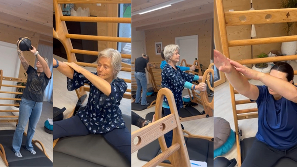 Drei Personen trainieren in einem Raum mit einem Reformer. Eine Person hält eine schwarze Kugel beim Dehnen. Eine andere Person dehnt sich im Sitzen, und eine dritte Person steht im Hintergrund.