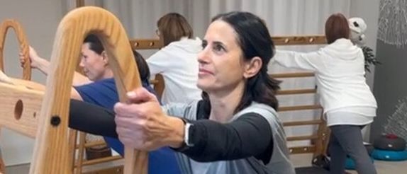 Eine Frau macht ein Training mit einem Holzwerkzeug. Sie steht auf einer Matte und scheint sich zu dehnen. Hinter ihr machen andere Leute Übungen. Sie tragen alle Turnschuhe. Sie sind in einem Raum mit einer Holzstange und einer Pflanze in der Ecke.