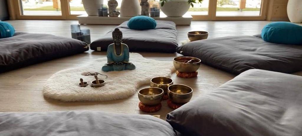 Ein ruhiger Meditationsraum mit grauen Kissen, einem weißen Fellteppich und einer sitzenden Buddha-Statue, umgeben von Schalen und Kerzen.