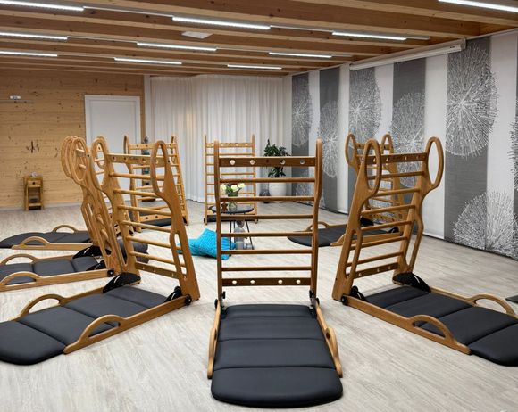 Ein Raum mit Holzwänden und -decke verfügt über mehrere Holz-Trainingsgeräte. Es gibt Liegestühle mit schwarzen Kissen. Ein Topf steht auf einem Tisch in der Nähe der Wand.