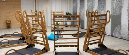 Ein Raum mit Holzwänden und -decke verfügt über mehrere Holz-Trainingsgeräte. Es gibt Liegestühle mit schwarzen Kissen. Ein Topf steht auf einem Tisch in der Nähe der Wand.