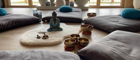 Ein ruhiger Raum mit grauen Kissen, einem weißen Teppich und einer Buddha-Statue. Schüsseln und andere Gegenstände sind auf dem Teppich platziert.