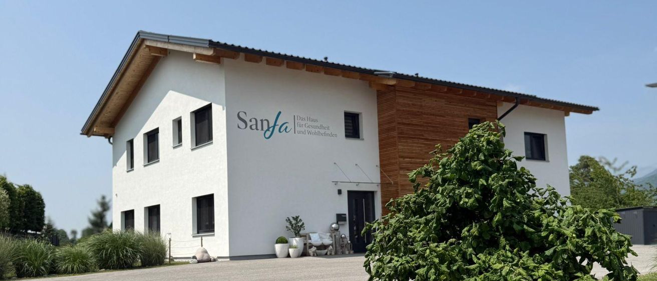 Ein weißes Gebäude mit einem braunen Dach, dem Wort 'Sanfa' auf der Vorderseite und mehreren Fenstern. Stühle stehen draußen und ein Busch ist davor.