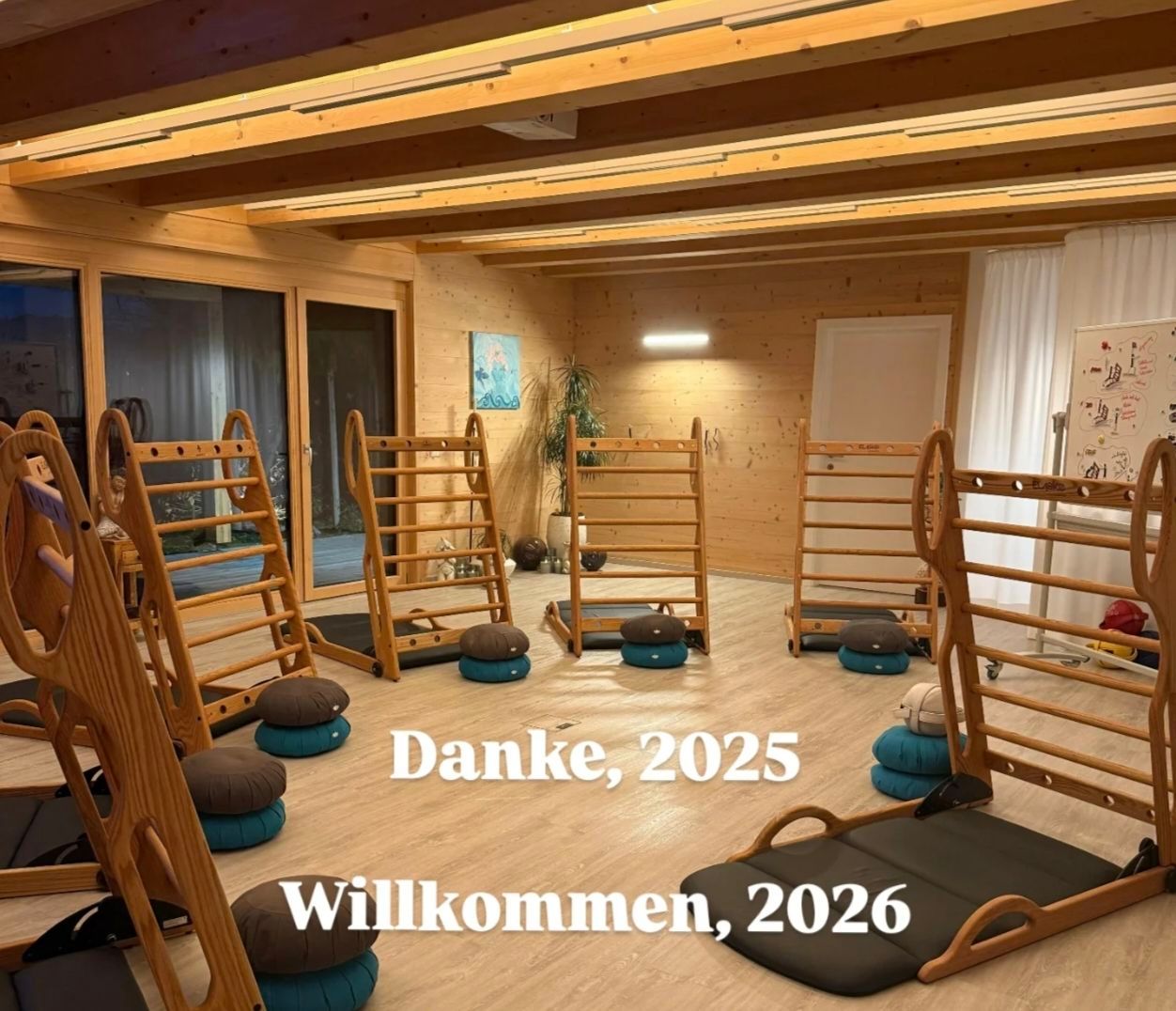 Ein Innenraum mit Holzböden und hölzernen Übungsgeräten, einschließlich Leitern und Kissen. Der Raum hat Holzwände und Deckenleuchten. Es gibt eine Pflanze und einen Bilderrahmen an der Wand. Der Text Danke, 2025 Willkommen, 2026 wird angezeigt.