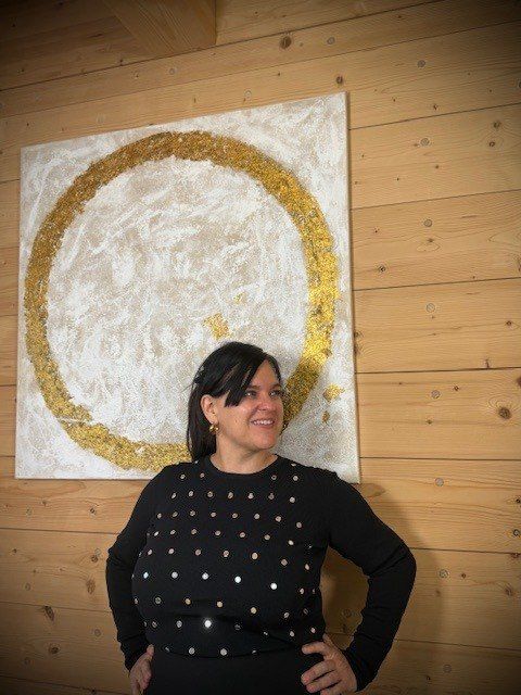 Eine Frau steht vor einem großen kreisförmigen Gemälde und lächelt für ein Foto. Das Gemälde zeigt einen goldenen Kreis auf weißem Hintergrund.