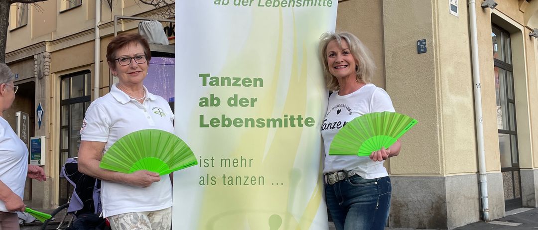 Zwei Frauen stehen neben einem Banner mit der Aufschrift 'Tanzen ab der Lebensmittel'. Sie halten grüne Fächer und lächeln für die Kamera. Ein Gebäude mit einem Schild, auf dem 'Miller' steht, ist im Hintergrund zu sehen.