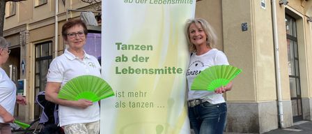 Zwei Frauen stehen neben einem Banner mit der Aufschrift 'Tanzen ab der Lebensmittel'. Sie halten grüne Fächer und lächeln für die Kamera. Ein Gebäude mit einem Schild, auf dem 'Miller' steht, ist im Hintergrund zu sehen.