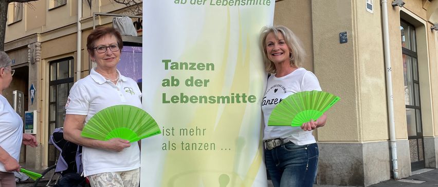 Zwei Frauen stehen neben einem Banner mit der Aufschrift 'Tanzen ab der Lebensmittel'. Sie halten grüne Fächer und lächeln für die Kamera. Ein Gebäude mit einem Schild, auf dem 'Miller' steht, ist im Hintergrund zu sehen.