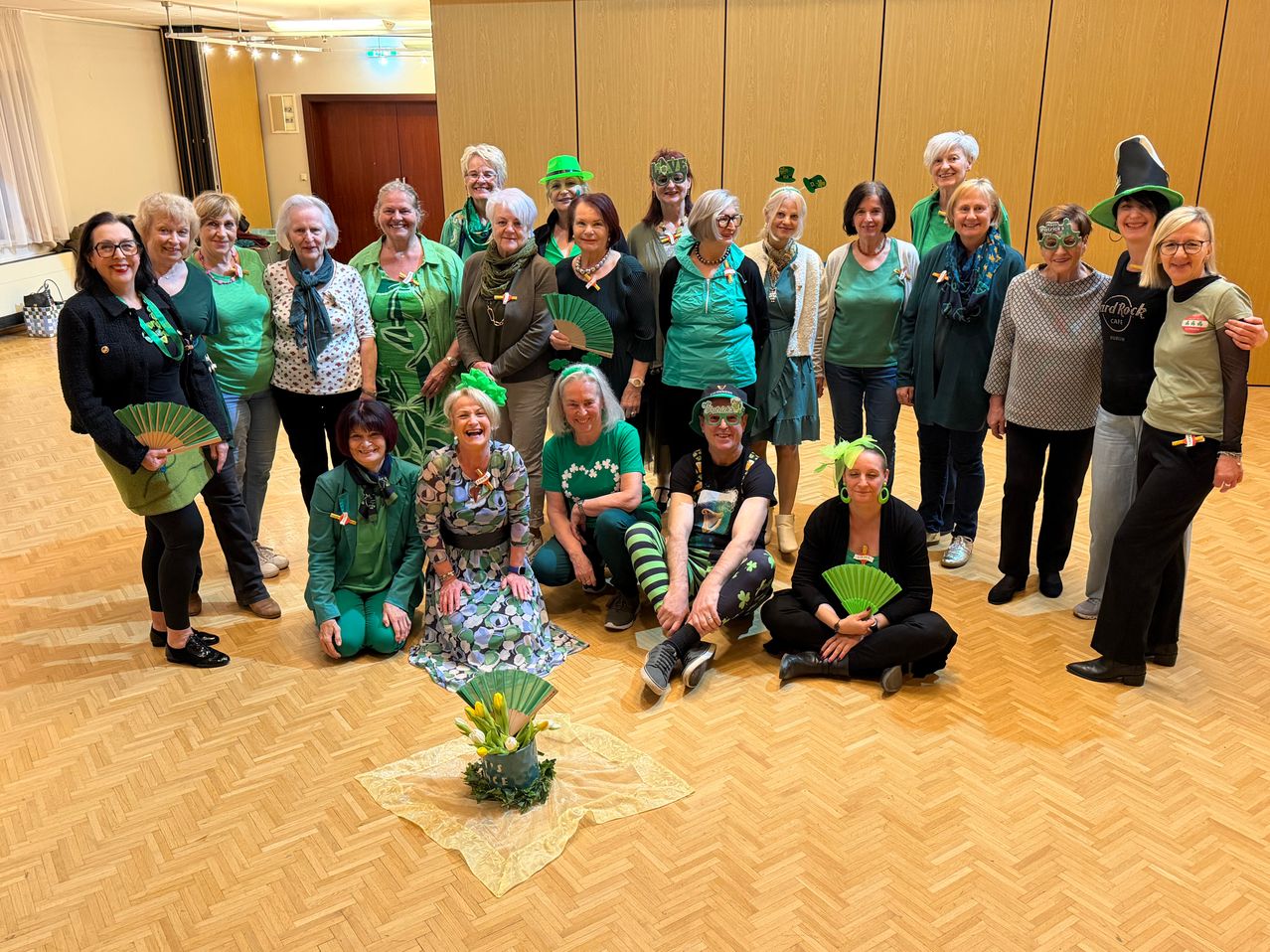 Eine Gruppe von Menschen in grüner Kleidung und Accessoires posiert für ein Foto in einem Raum, einige sitzend und andere stehend, mit einem Holzboden und einer Wand dahinter.