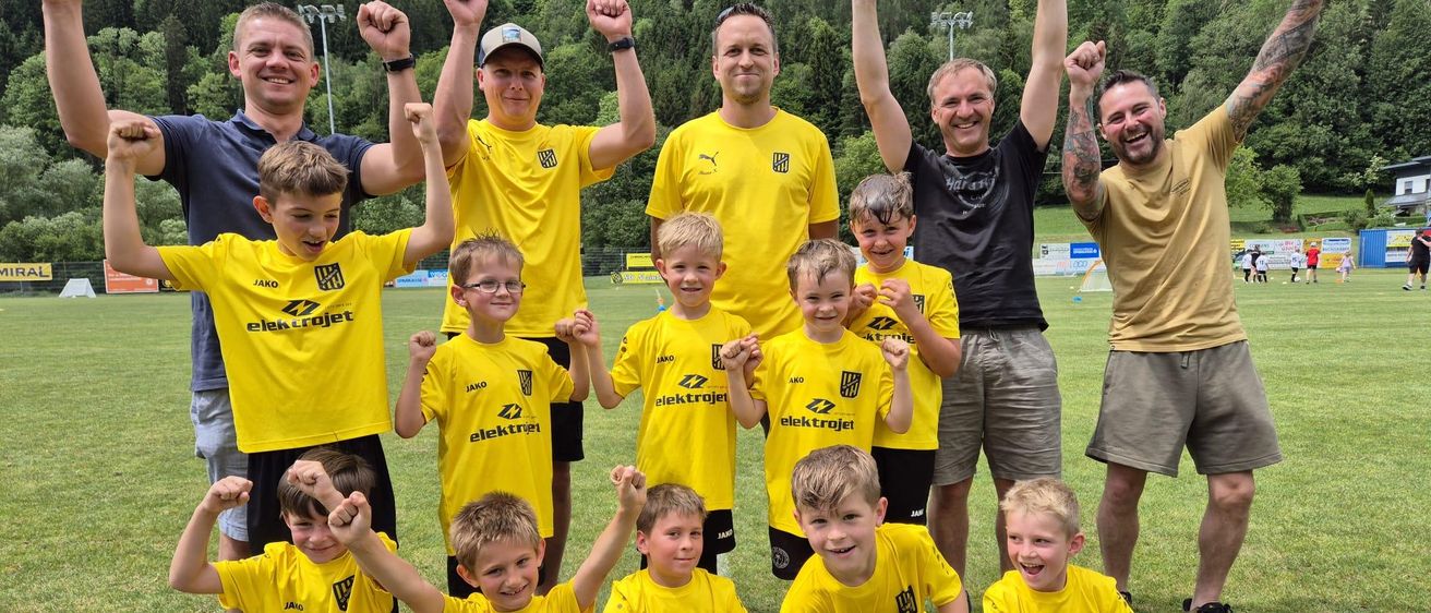 Eine Gruppe junger Jungen in gelben und schwarzen Fußballtrikots posiert für ein Foto mit ihren Trainern auf einem Rasenfeld mit einer malerischen Kulisse.