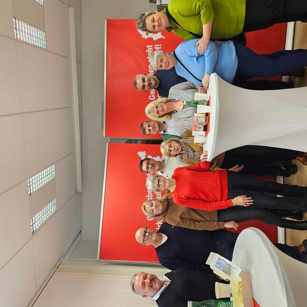 Eine Gruppe von Menschen steht vor einem roten Banner mit Essen und Getränken auf einem Tisch. Sie lächeln für ein Foto.