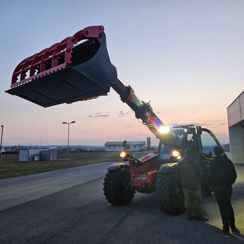 A red JCB telehandler is parked on the road with two people observing, under a sunset sky.