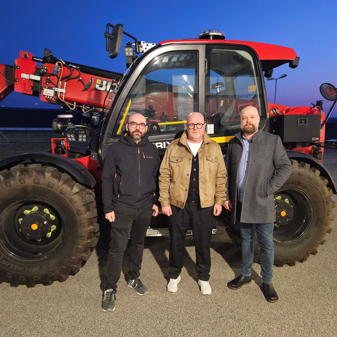 Three men stand in front of a large red tractor. The man on the left wears glasses and a black jacket. The middle man wears a brown jacket, and the man on the right wears a gray jacket. The tractor has a large front tire and a red cab.