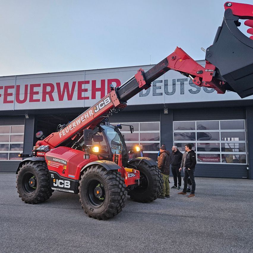 A red JCB telehandler with a black bucket is parked in front of a building labeled 'Feuerwehr'. Four men are standing around it, two of them wearing hats.