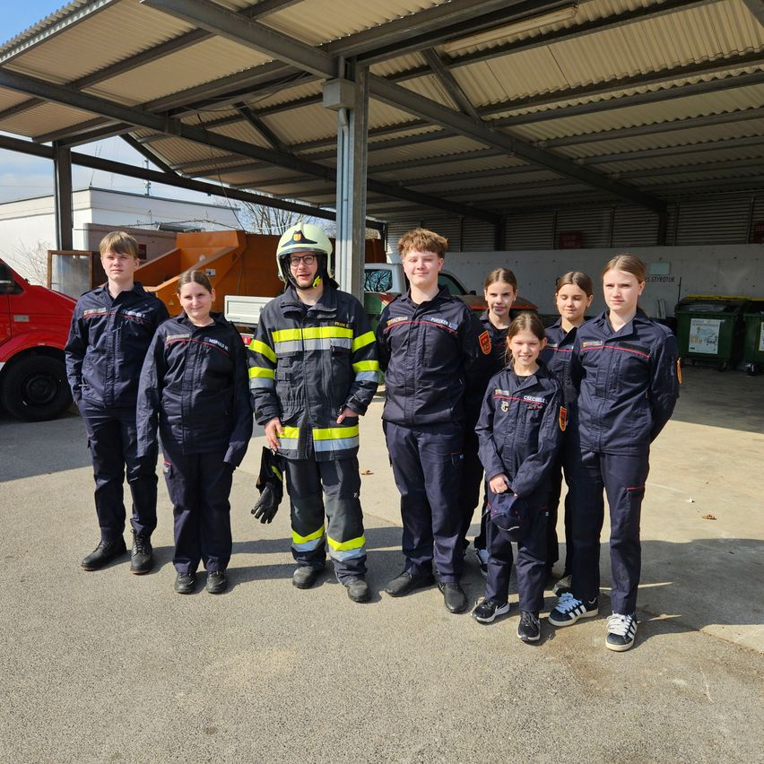 Eine Gruppe junger Menschen in Uniform, einige mit Helmen, stehen in einer Garage. Ein Mann in der Mitte hält einen Handschuh. Ein roter Lkw und Mülleimer im Hintergrund.