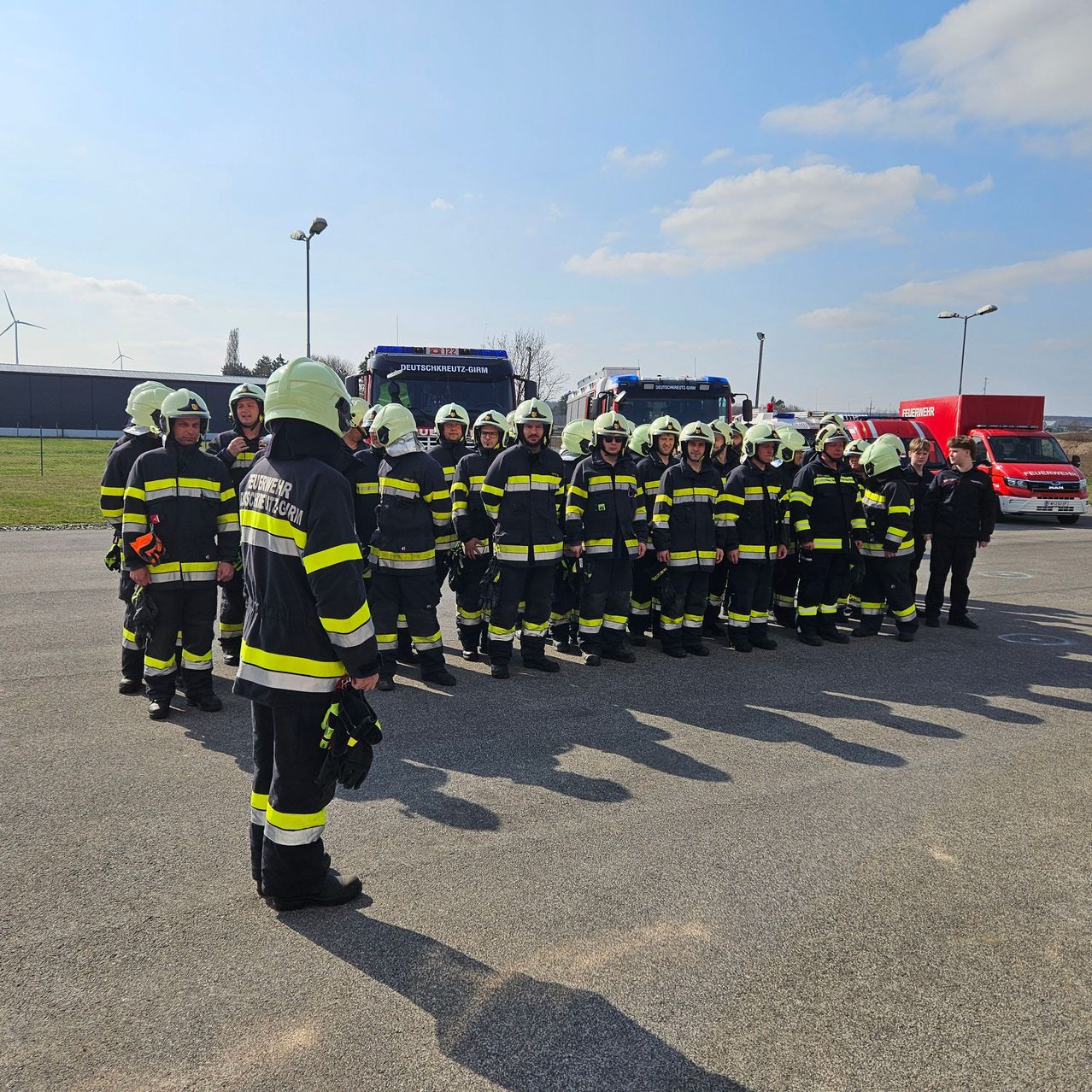 Eine Reihe von Feuerwehrleuten in Uniform und Helmen steht in einem Freiluftbereich mit Notfallfahrzeugen im Hintergrund.