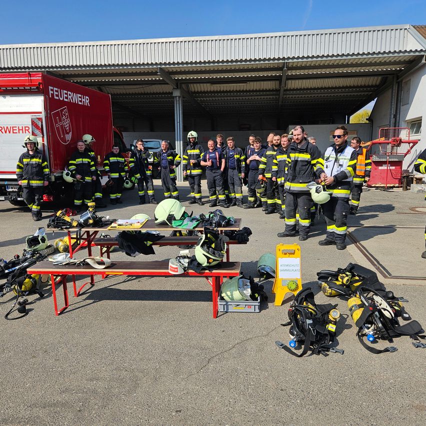 Feuerwehrleute in Uniform stehen vor einer Feuerwache in einer Reihe. Ausrüstung und Tische sind draußen aufgebaut. Ein Feuerwehrauto ist in der Nähe geparkt.
