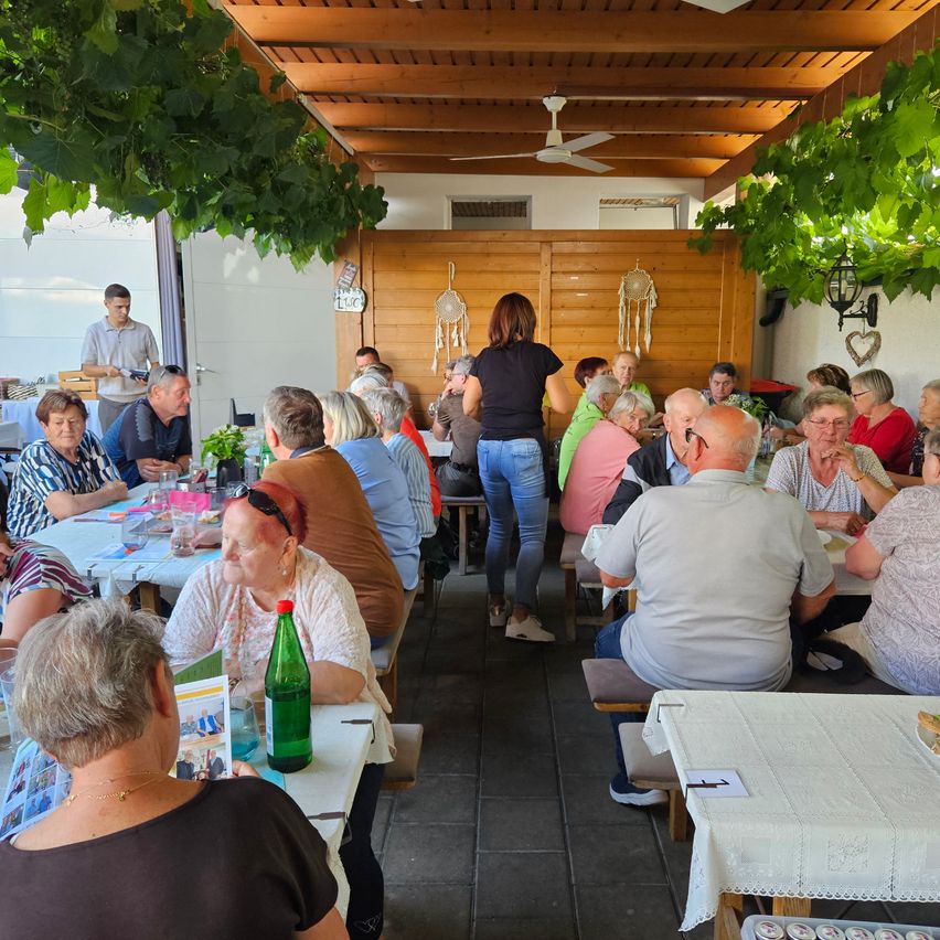 Eine Gruppe älterer Menschen diniert in einem Restaurant mit Holzwänden und einem Deckenventilator. Einige sitzen an Tischen, während andere stehen und sich unterhalten.