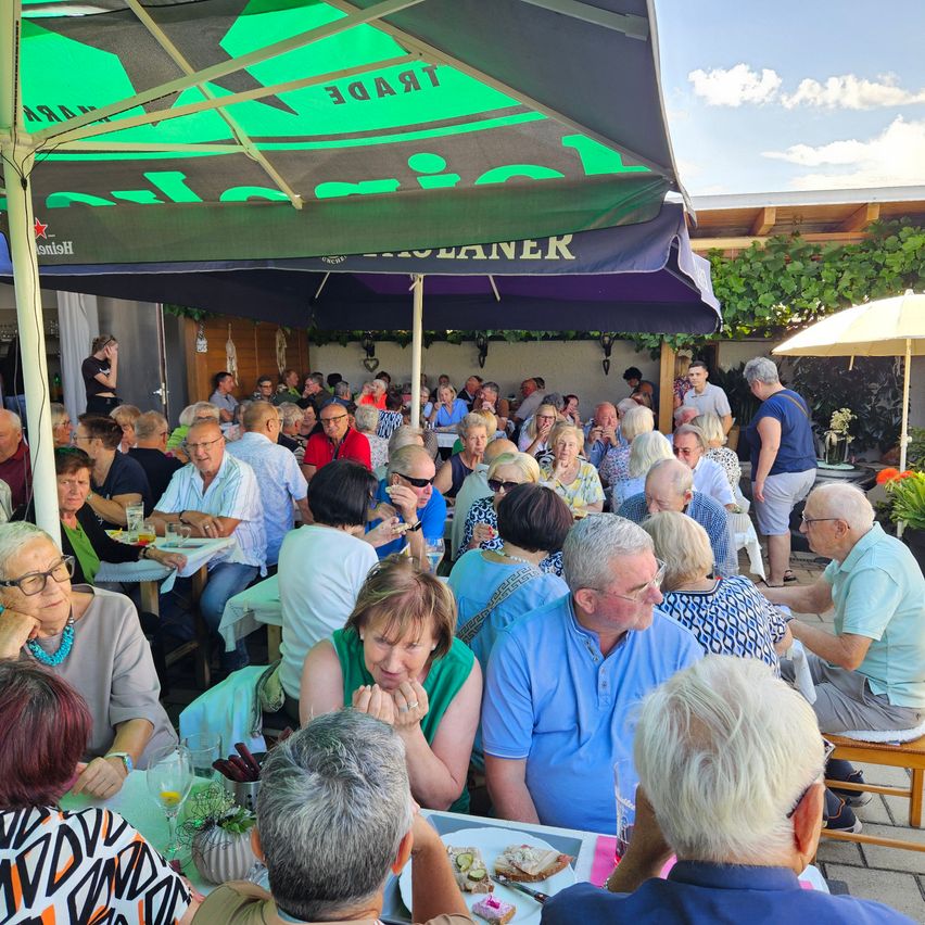 Eine Gruppe älterer Menschen isst im Freien unter einem großen grünen Sonnenschirm. Sie sitzen an Tischen, einige stehen, und scheinen ihre Mahlzeit zu genießen. Die Umgebung ist im Freien mit klarem Himmel und etwas Grün im Hintergrund.