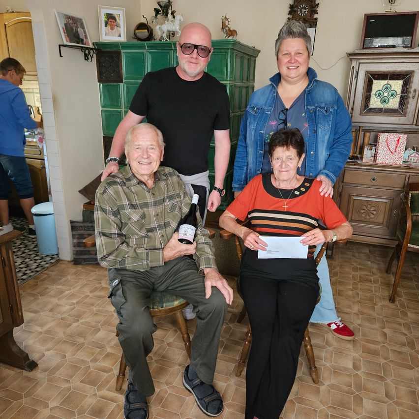Three people are sitting in a room, two of them are smiling and one is holding a bottle of wine. The woman is holding a piece of paper. Behind them is a cabinet with a clock on top.