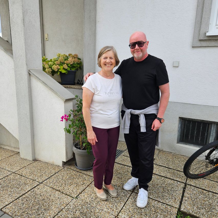 A man and a woman stand together in front of a house, smiling and posing for a photo. The man wears sunglasses and a black shirt, while the woman wears a white shirt and purple pants. Behind them, there are potted plants and a bicycle.