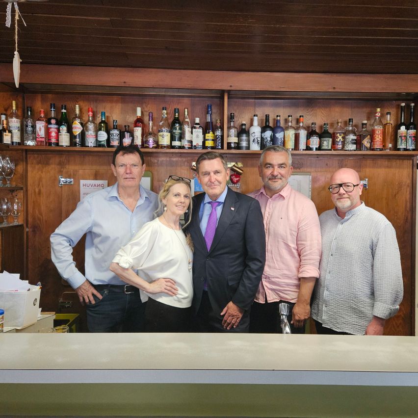 Five people stand behind a bar counter, smiling for a photo. They are dressed in casual to formal attire. Behind them, a wooden shelf holds various bottles of liquor.