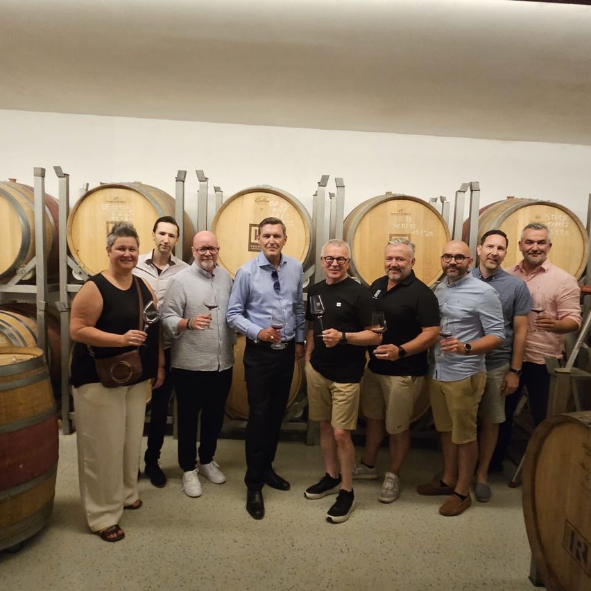 A group of people in a wine cellar, holding wine glasses and smiling for a photo. Large wooden barrels are stacked behind them.