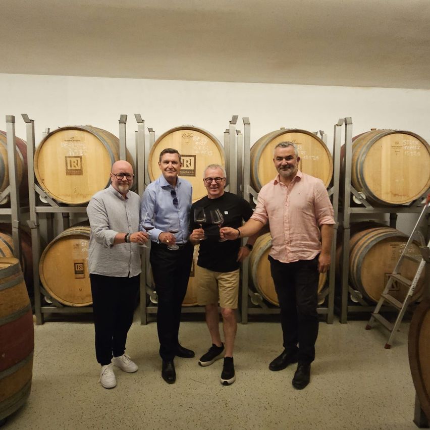 Four men in a wine cellar, holding wine glasses, smiling and posing for a photo. Behind them, large wooden barrels are stacked on metal racks.