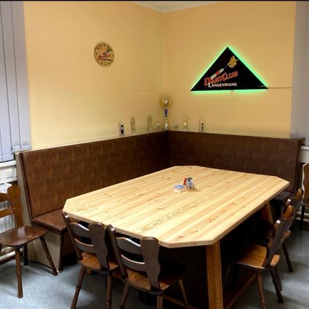 Ein Essbereich mit einem Holztisch und Stühlen, einer Eckbank und Trophäen an der Wand. Ein beleuchtetes Dartboard-Schild ist an der Wand angebracht.