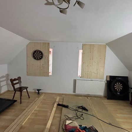 Ein Dachbodenzimmer mit Holzwänden und -boden, zwei kleinen Fenstern und einem Dartboard an einer Wand. Ein Holzstuhl, ein Heizkörper und einige Werkzeuge sind ebenfalls zu sehen.