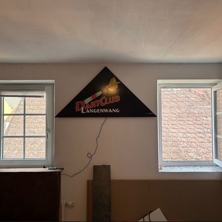 Ein Schild für den Dart Club Langenwang ist an einer Wand angebracht. Zwei Fenster befinden sich auf beiden Seiten des Schildes. Ein Heizkörper ist unter dem Schild.