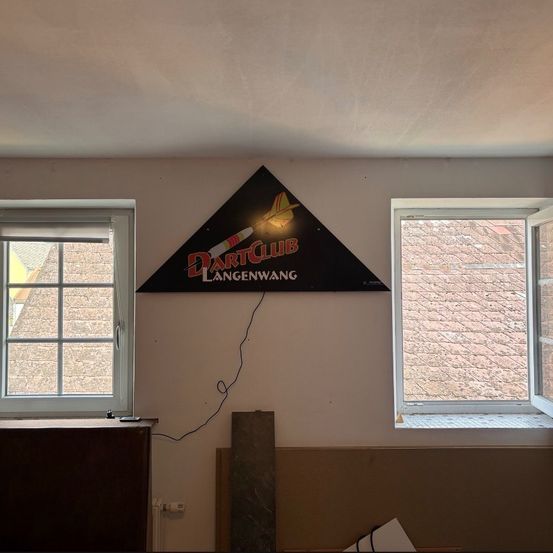 Ein Schild für den Dart Club Langenwang ist an einer Wand angebracht. Zwei Fenster befinden sich auf beiden Seiten des Schildes. Ein Heizkörper ist unter dem Schild.