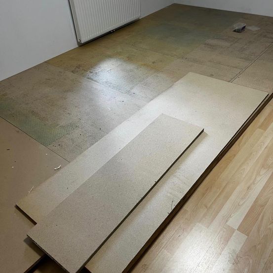 Ein leerer Raum mit Holzboden und Pappblättern. An der Wand ist ein weißer Heizkörper angebracht.