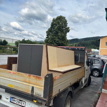 Ein Lastwagen mit dem Kennzeichen BM 294 EY steht auf einem Parkplatz mit Schränken und Holzbrettern im Heck. In der Nähe ist ein Auto geparkt, und im Hintergrund ist ein Baum zu sehen.