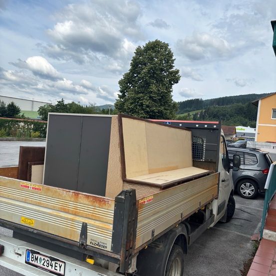 Ein Lastwagen mit dem Kennzeichen BM 294 EY steht auf einem Parkplatz mit Schränken und Holzbrettern im Heck. In der Nähe ist ein Auto geparkt, und im Hintergrund ist ein Baum zu sehen.