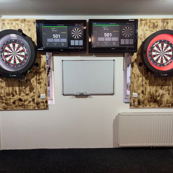 Drei Dartscheiben sind an einer hölzernen Wand mit weißem Hintergrund montiert. Zwei digitale Bildschirme zeigen die Punktzahl. Ein Bildschirm zeigt 501 an. Ein Whiteboard ist unterhalb der Bildschirme montiert.