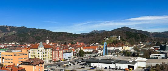 Luftaufnahme einer Stadt mit einer Mischung aus modernen und traditionellen Gebäuden. Die Stadt ist von Hügeln und Bergen umgeben. Der Himmel ist blau mit einigen Wolken.