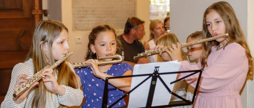 Vier Mädchen spielen Flöten in einem Raum. Sie schauen auf Musikblätter auf einem Ständer. Im Hintergrund beobachten ein Mann und zwei Frauen sie.