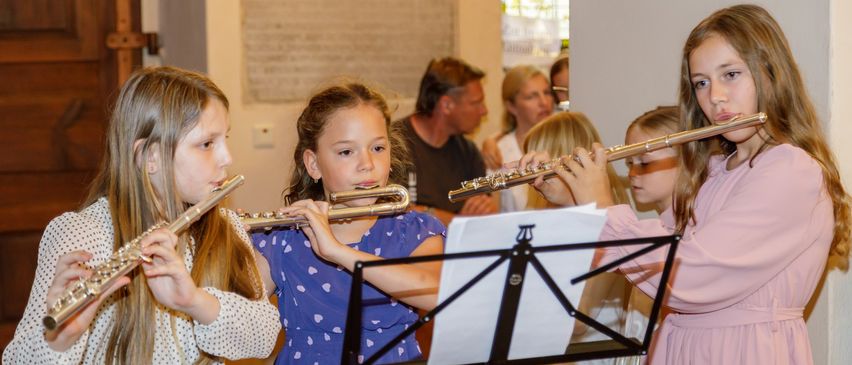 Vier Mädchen spielen Flöten in einem Raum. Sie schauen auf Musikblätter auf einem Ständer. Im Hintergrund beobachten ein Mann und zwei Frauen sie.