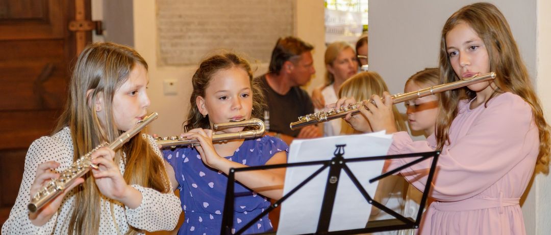 Vier Mädchen spielen Flöten in einem Raum. Sie schauen auf Musikblätter auf einem Ständer. Im Hintergrund beobachten ein Mann und zwei Frauen sie.