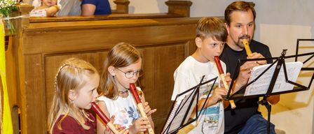 Ein Mann und drei Kinder spielen Blockflöten in einer Kirche, mit Musikblättern vor ihnen.