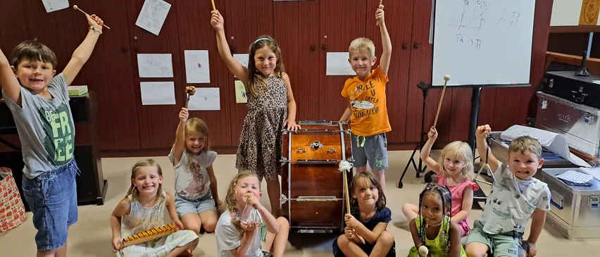 Eine Gruppe von Kindern in einem Klassenzimmer, die Musikinstrumente halten und lächeln. Eine Trommel steht in der Mitte, und ein Whiteboard ist im Hintergrund sichtbar.