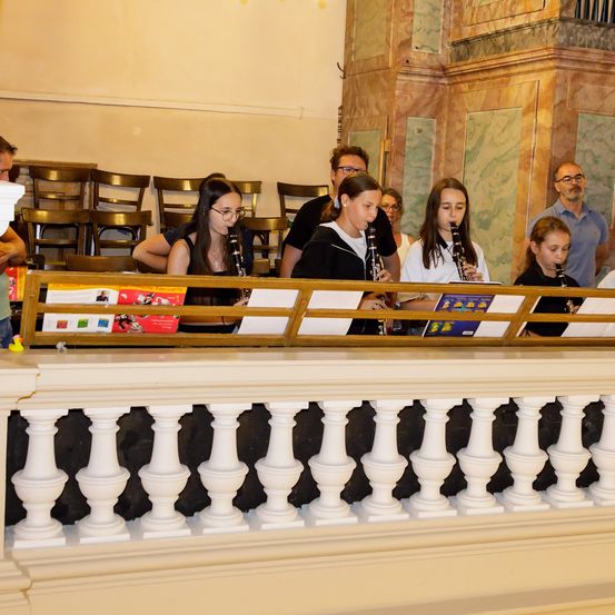 Eine Gruppe junger Mädchen spielt Klarinetten auf einem Balkon. Dahinter beobachten Erwachsene. Der Balkon hat weiße Geländer.